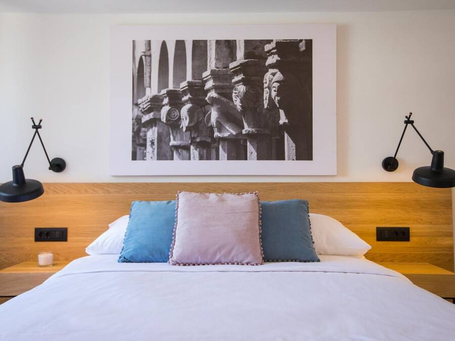 Een net opgemaakt bed met een houten hoofdbord, twee bedlampjes en een zwart-wit foto in een lijst aan de muur.