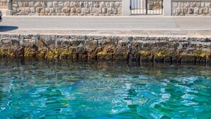 Un bâtiment en pierre doté de nombreuses fenêtres et volets, une entrée fermée par un portail et une vue dégagée sur l’eau au premier plan.