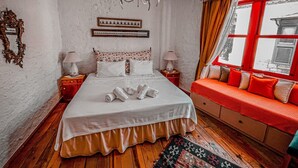 A bedroom with a bed, bedside tables, a chandelier, a window with red curtains, and a view of a building outside. - Narcicegi Alacati (Cesme)