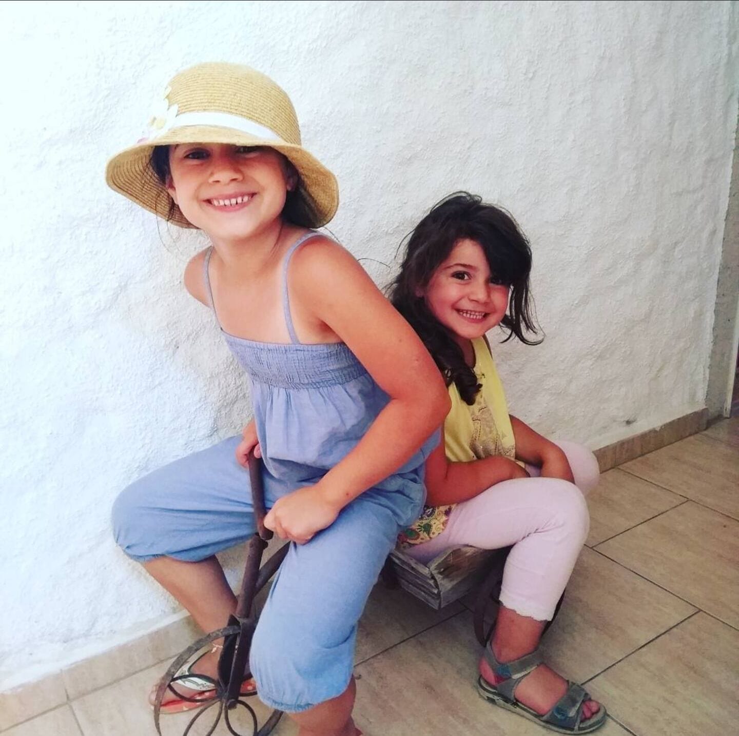 Deux enfants sont assis sur un banc, adossés à un mur uni.