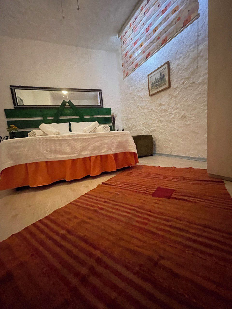 A room with a bed, a green headboard, a white wall, a framed picture, and a patterned rug.