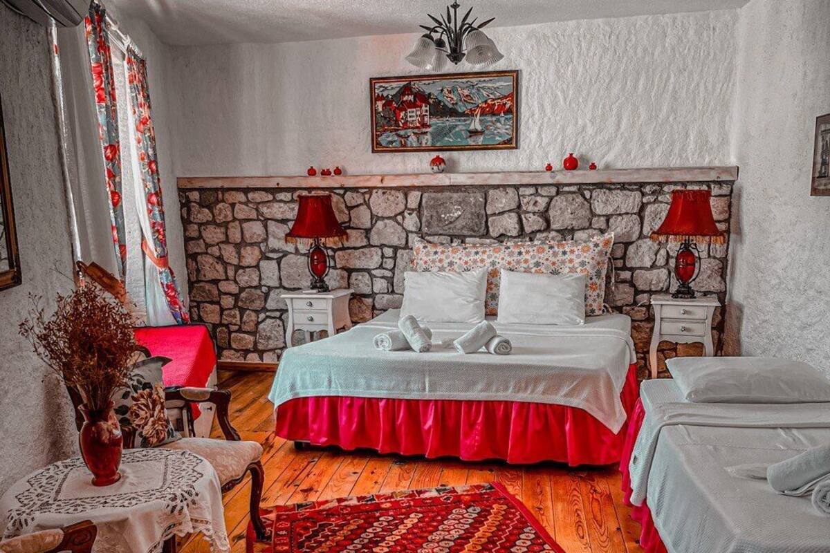 A bedroom with a stone wall, two beds with red bedding, a wooden floor, a red patterned rug, and a painting above the bed.