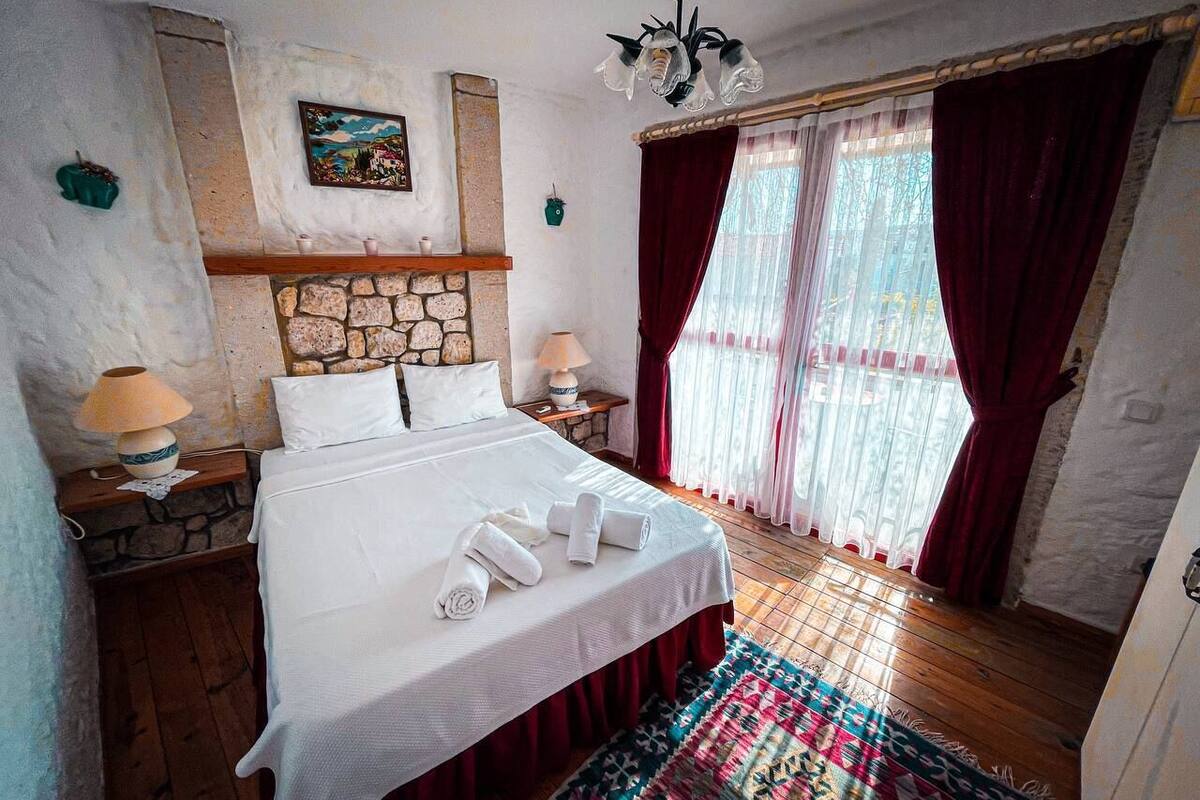 A bedroom with a stone fireplace, a bed with white linens, a chandelier, and a view through sheer curtains.