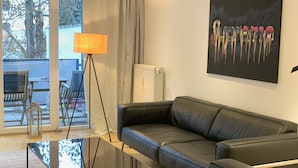 Una sala de estar moderna con un sofá de cuero negro, una mesa de centro de vidrio y una lámpara de pie. Se observa un ventanal con cortinas, un radiador y un cuadro en la pared.