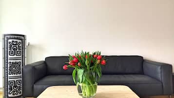 Un sofá de cuero negro frente a una pared blanca, con un panel decorativo blanco y negro a la izquierda. Delante del sofá, una mesa de centro de madera con un jarrón de vidrio transparente que contiene tulipanes rojos.