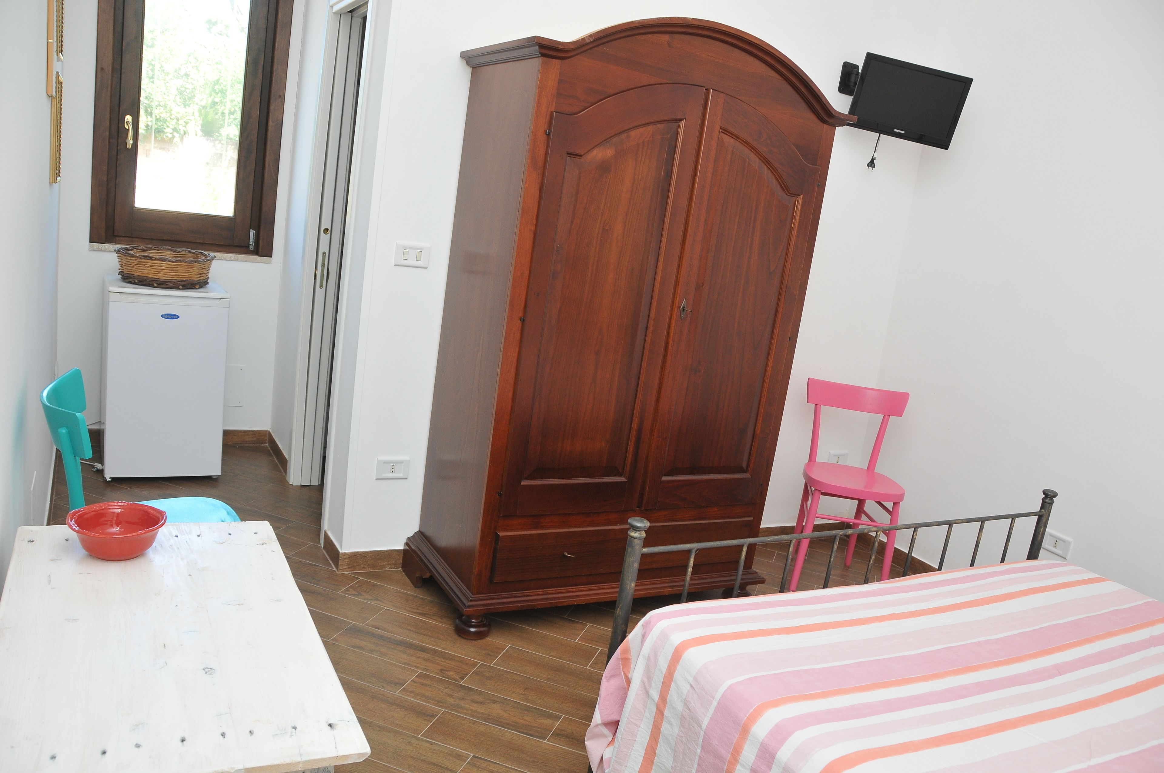 A bedroom with a bed, a wardrobe, a desk with a red bowl, a pink chair, and a television mounted on the wall.