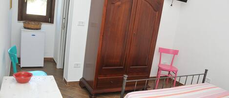 A bedroom with a bed, a wardrobe, a desk with a red bowl, a pink chair, and a television mounted on the wall.