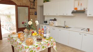 Cocina con armarios blancos, microondas y un lavabo. Hay una mesa con mantel floral y diversos alimentos.