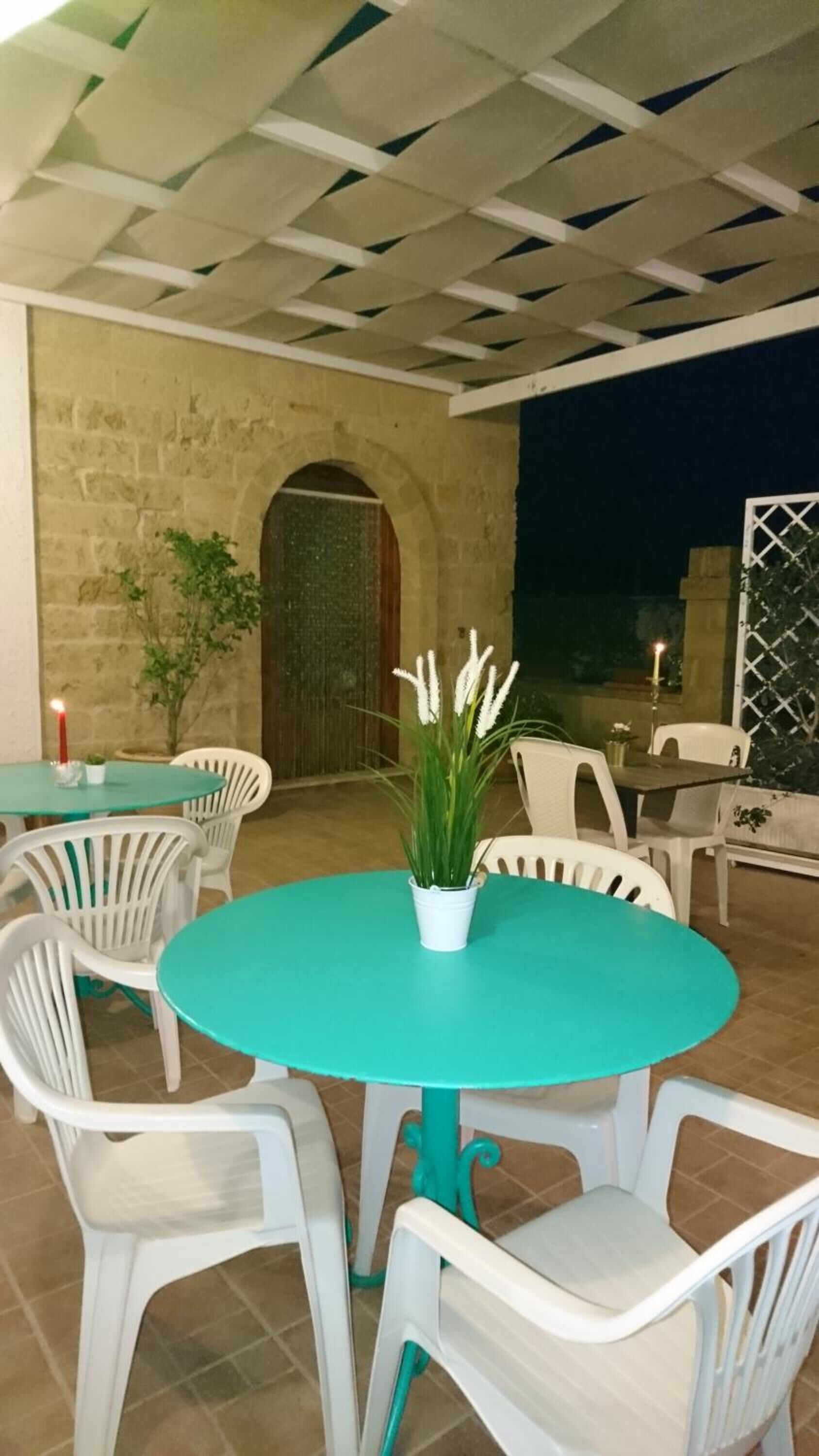 A round turquoise table with white plastic chairs, a potted plant, and a candle on a table in an outdoor patio area with stone walls and a wooden door.