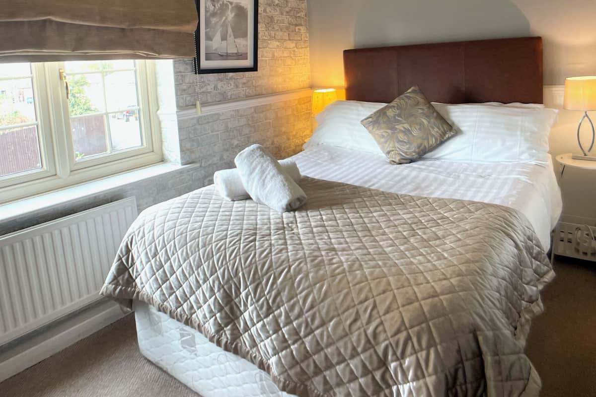 A neatly made bed with a quilted bedspread, a headboard, and two pillows. A framed picture hangs on the brick wall, and a window with blinds is visible.