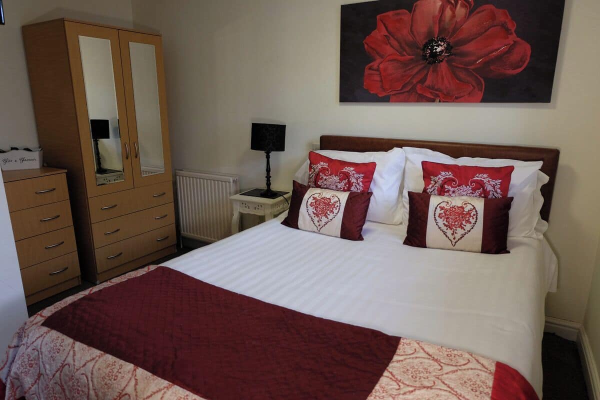 A bedroom with a bed, a wardrobe, a radiator, a lamp, and a large floral painting on the wall.