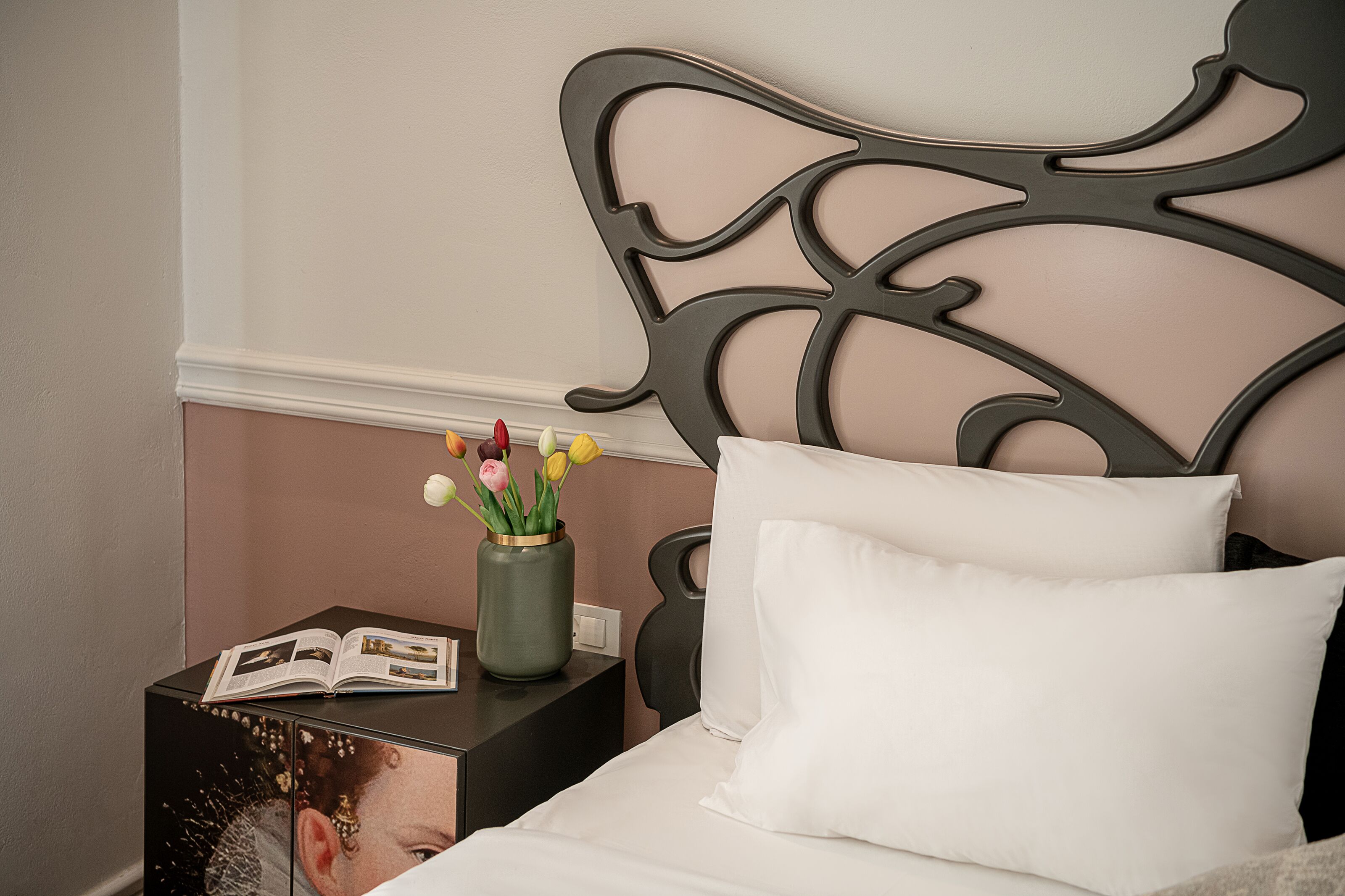 A neatly made bed with white pillows and a decorative headboard. A small table beside the bed holds a book and a vase with flowers.