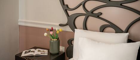 A neatly made bed with white pillows and a decorative headboard. A small table beside the bed holds a book and a vase with flowers.