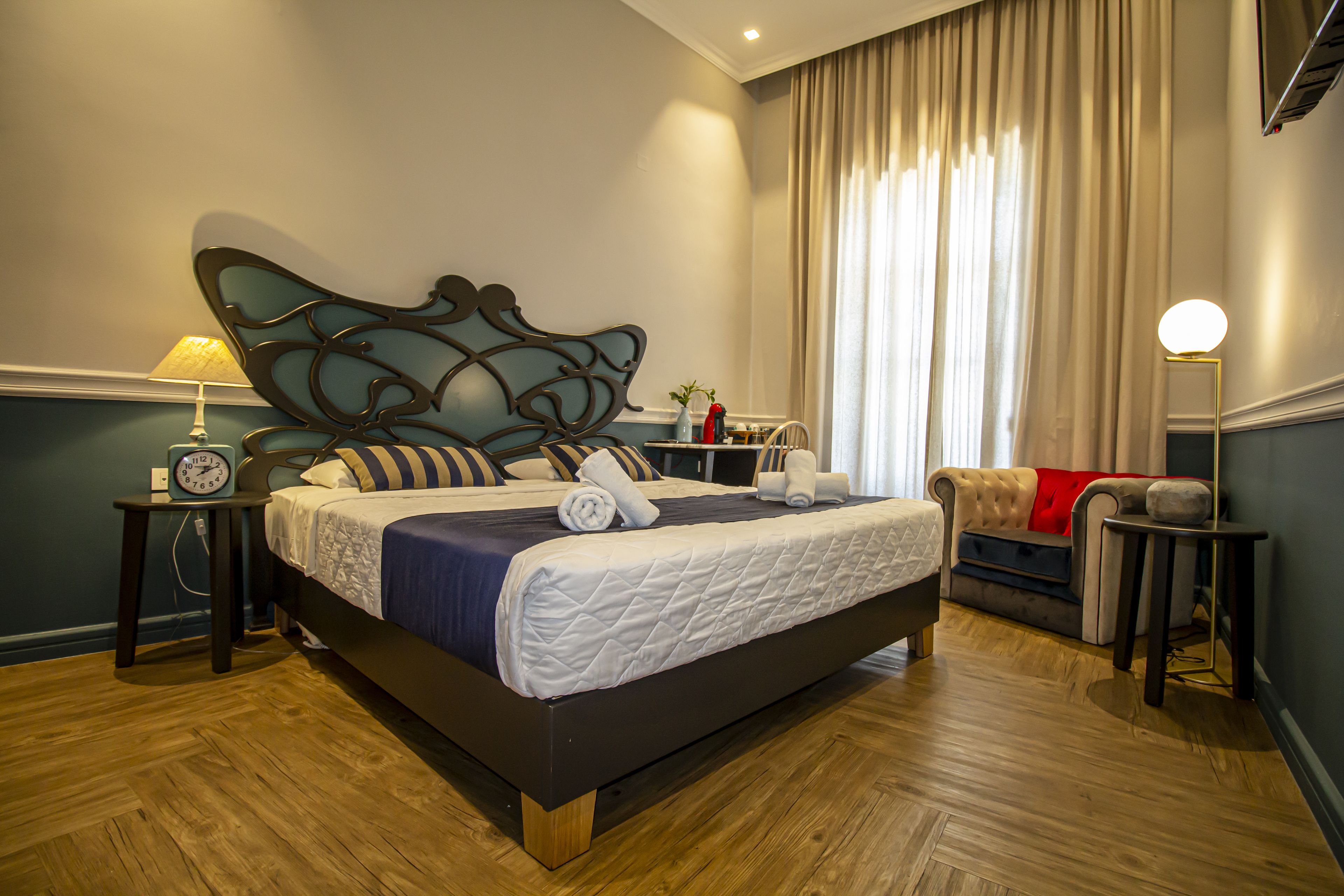 Ein ordentlich eingerichtetes Hotelzimmer mit einem großen Bett, einem Nachttisch mit Lampe, einer Uhr und einem Sofa mit roter Tagesdecke.