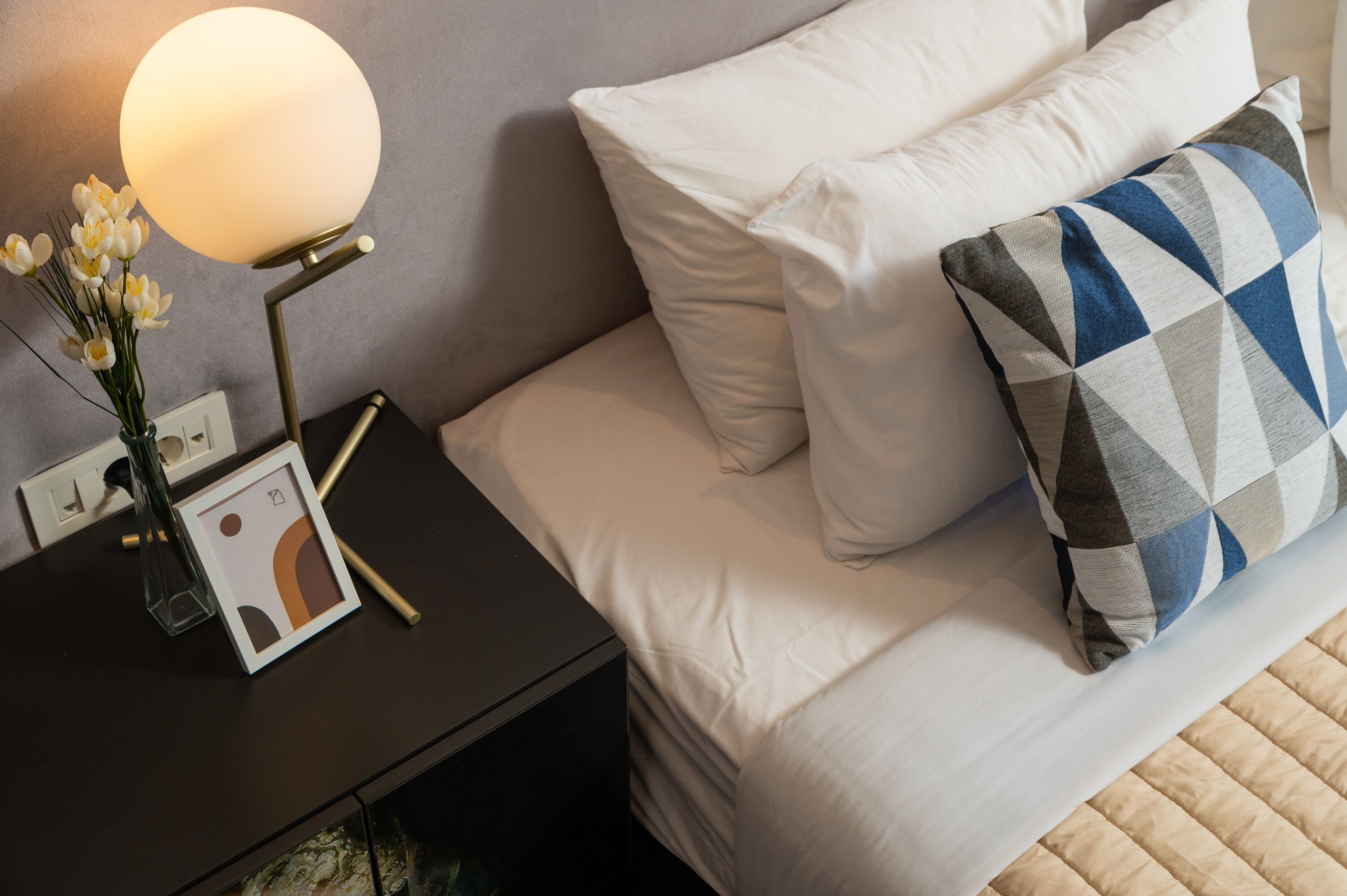 Een net opgemaakt bed met witte lakens en een kussen met dessin, een nachtkastje met een lamp en een vaas met bloemen.