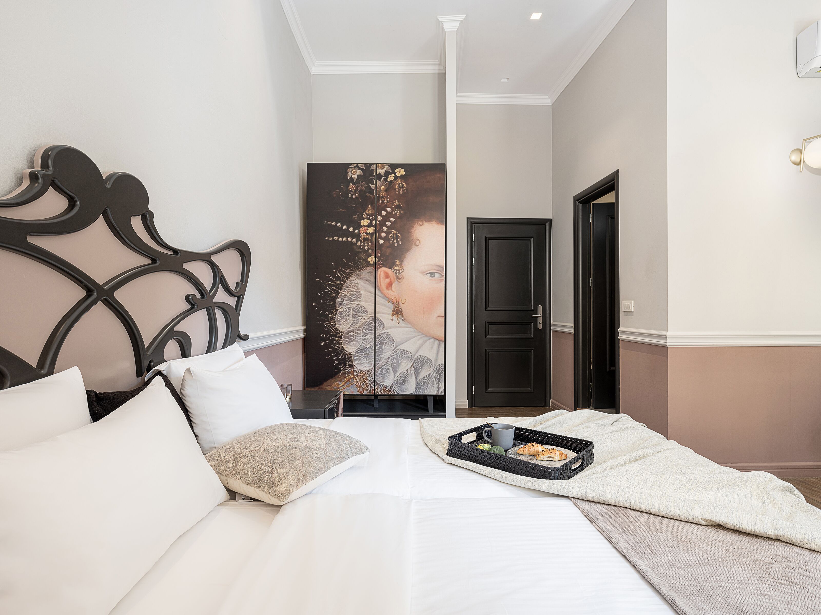 Un dormitorio con cama, almohadas, una bandeja con taza y bollería, un cuadro y una puerta.