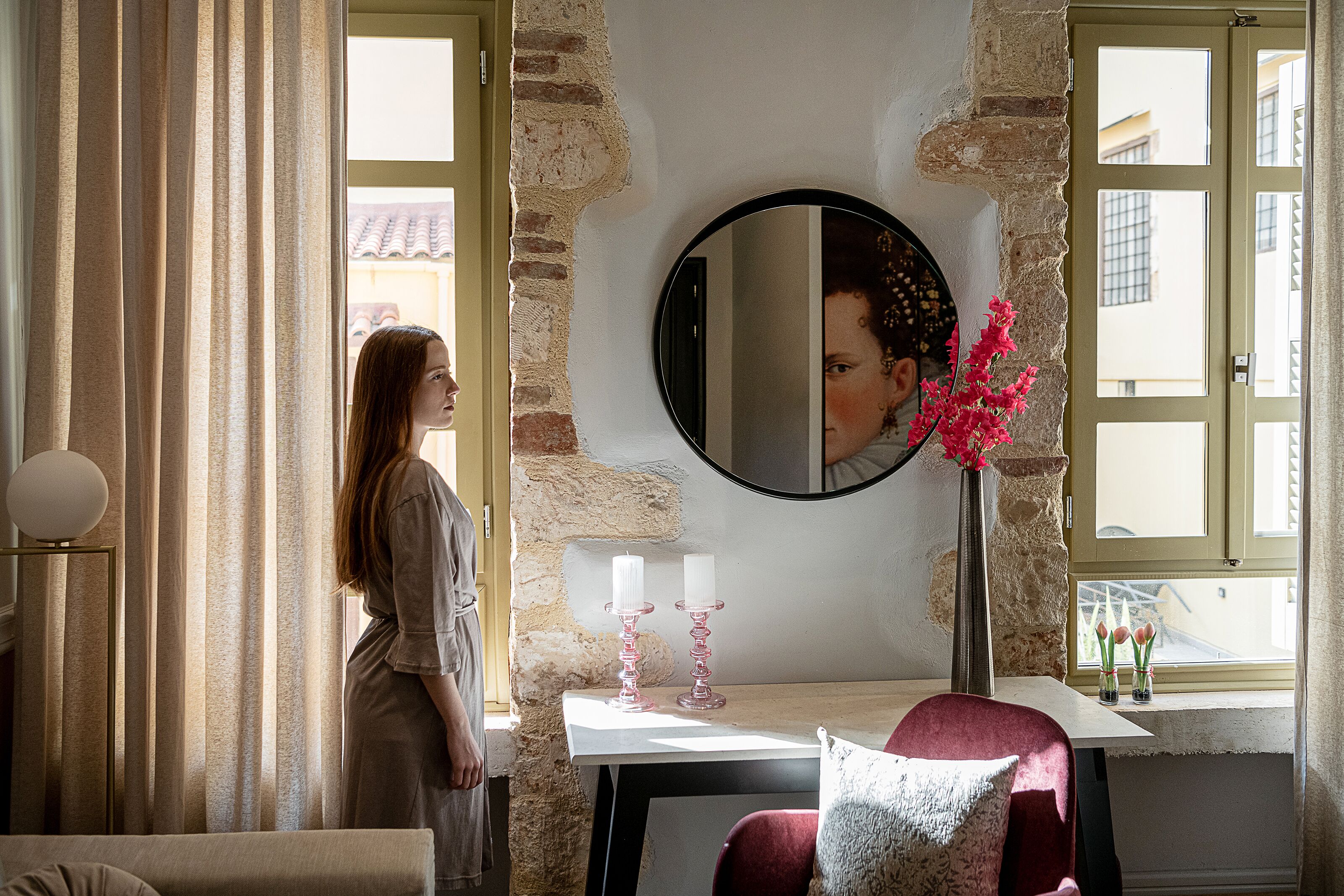 Una habitación con pared de piedra, un espejo redondo, una mesa con un jarrón de flores y una ventana con cortinas.