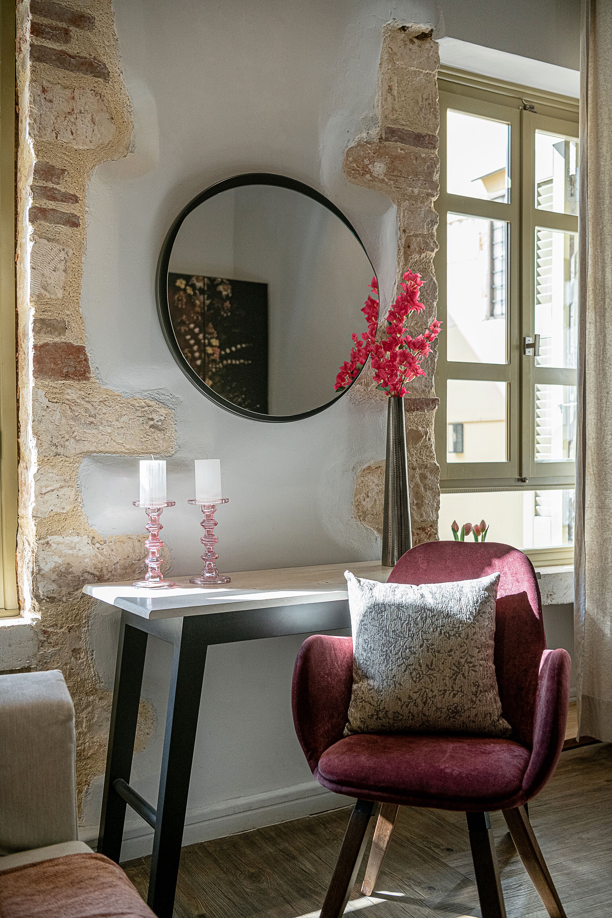 A room with a stone wall, a round mirror, a vase with flowers, a table with candles, and a red chair with a patterned pillow.