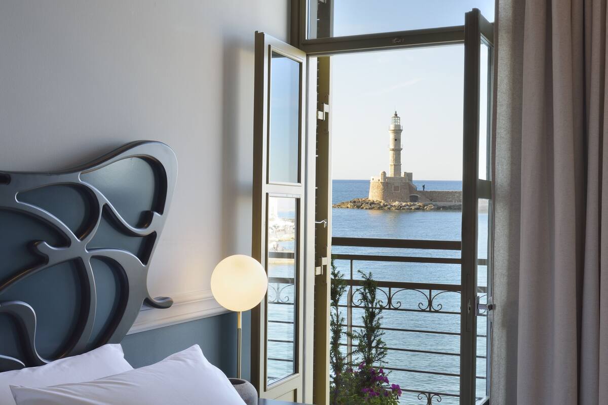 A hotel room with a bed, a nightstand, a lamp, and a view of a lighthouse through an open balcony door.