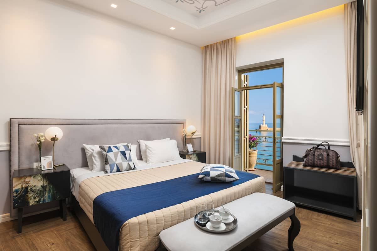 A hotel room with a large bed, bedside tables, a bench, and a view of the sea.