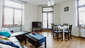 Ett modernt vardagsrum med en soffa, ett soffbord, ett matbord med stolar, en tv och stora fönster med gardiner.
