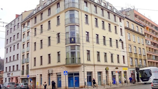 En byggnad med flera våningar, en entré i hörnet, flera butiker på bottenvåningen och ett tydligt markerat övergångsställe för fotgängare.