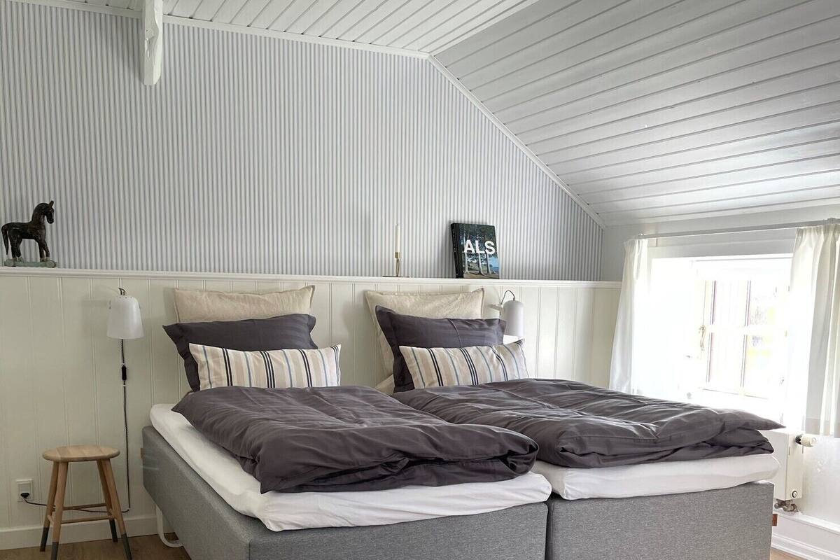 Un dormitorio moderno con techo estilo loft, una cama gris con almohadas a rayas, un taburete pequeño y un radiador.