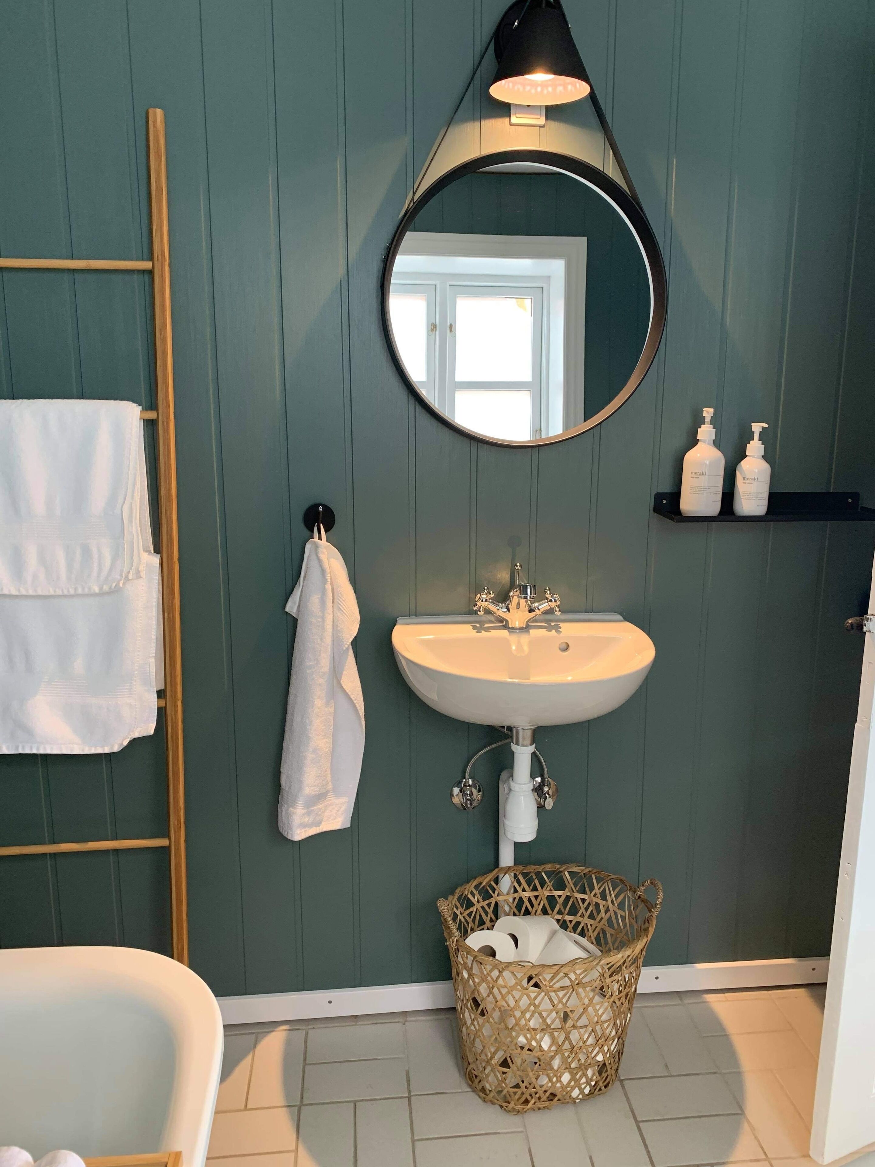 A bathroom with a white sink, a round mirror, a wooden ladder, a white bathtub, and a wicker basket.