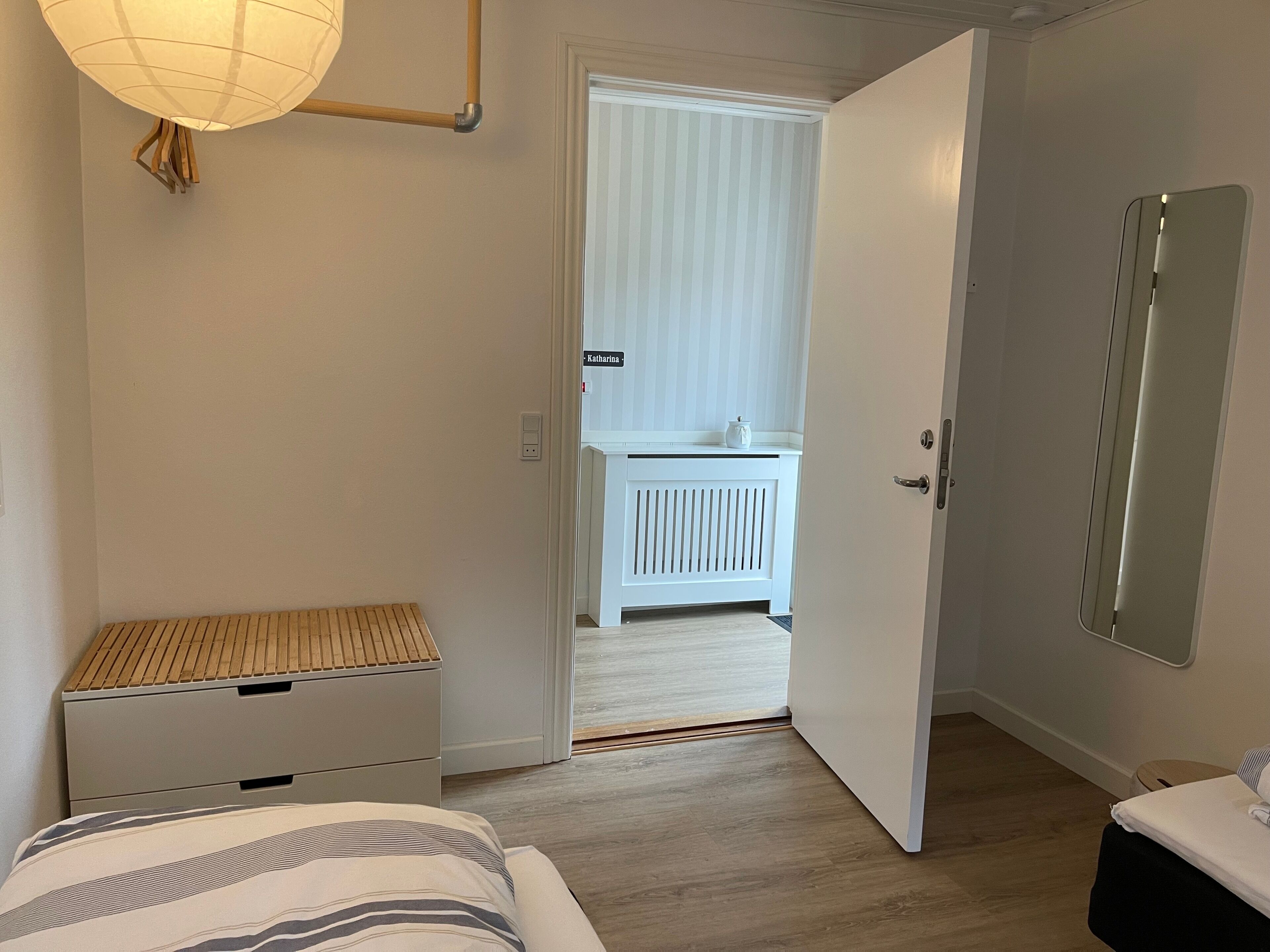 Un dormitorio con cama, mesita de noche y lámpara. Se ve una puerta que da a un baño con espejo e inodoro.