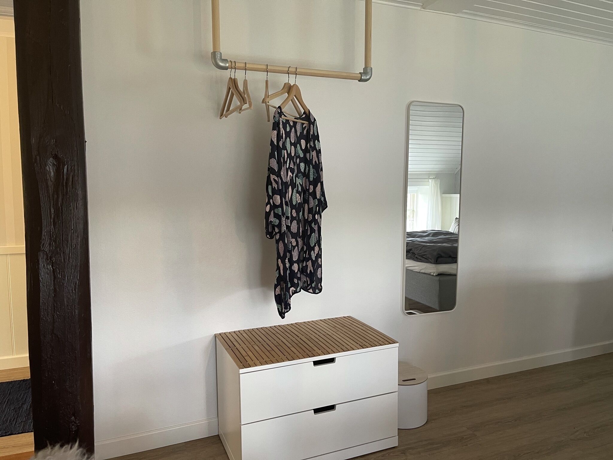 A bedroom with a wooden wardrobe, a hanging rack with clothes, a mirror, and a bedside table.