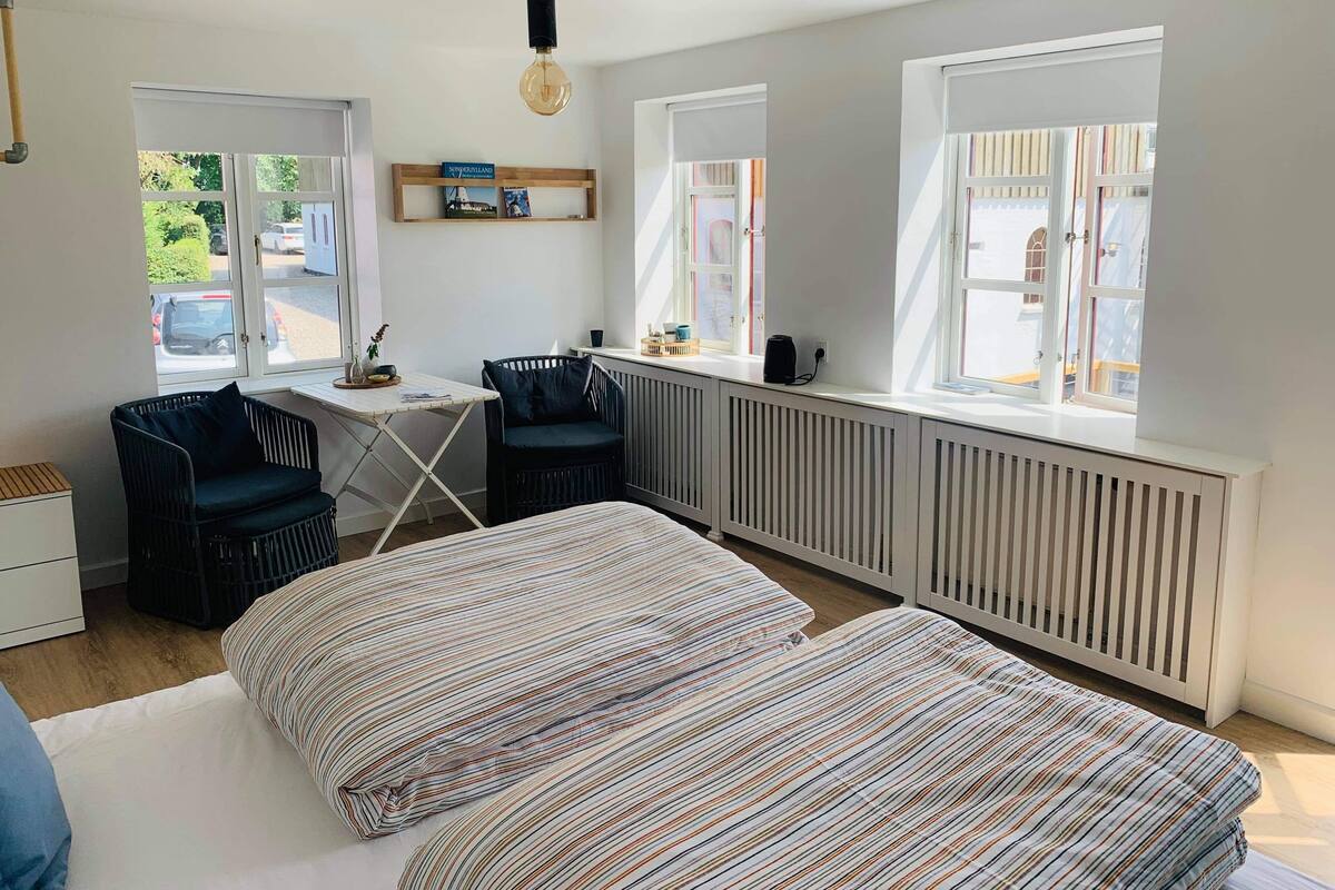 Una habitación con una cama, dos sillas, una mesita y un radiador. Se observan ventanas con persianas, un estante con libros y una lámpara colgante.