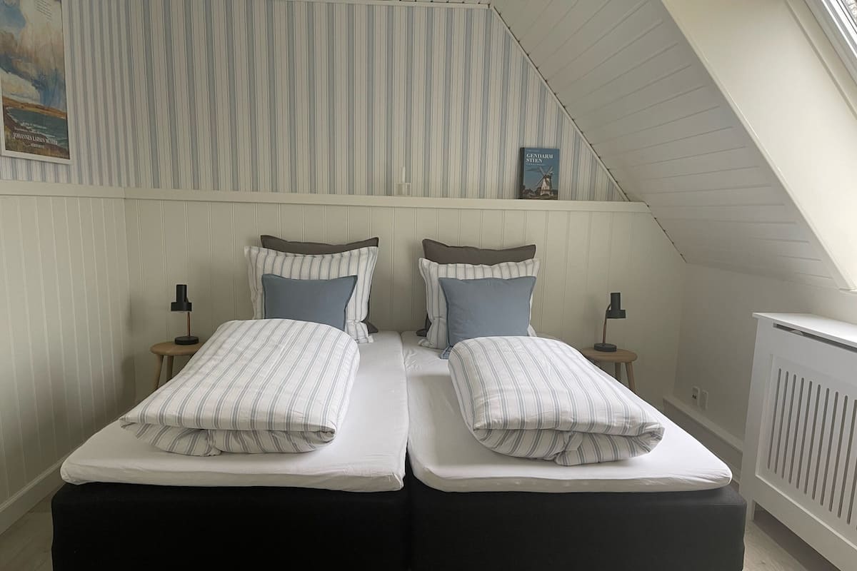 Habitación con dos camas individuales, ropa de cama a rayas, mesitas de noche con lámparas, un radiador y techo inclinado.