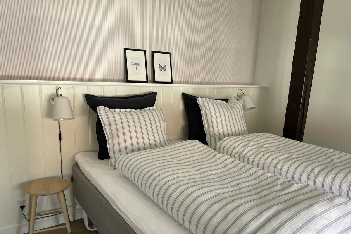 Una cama bien hecha con ropa de cama a rayas, una lámpara de noche y un taburete de madera.