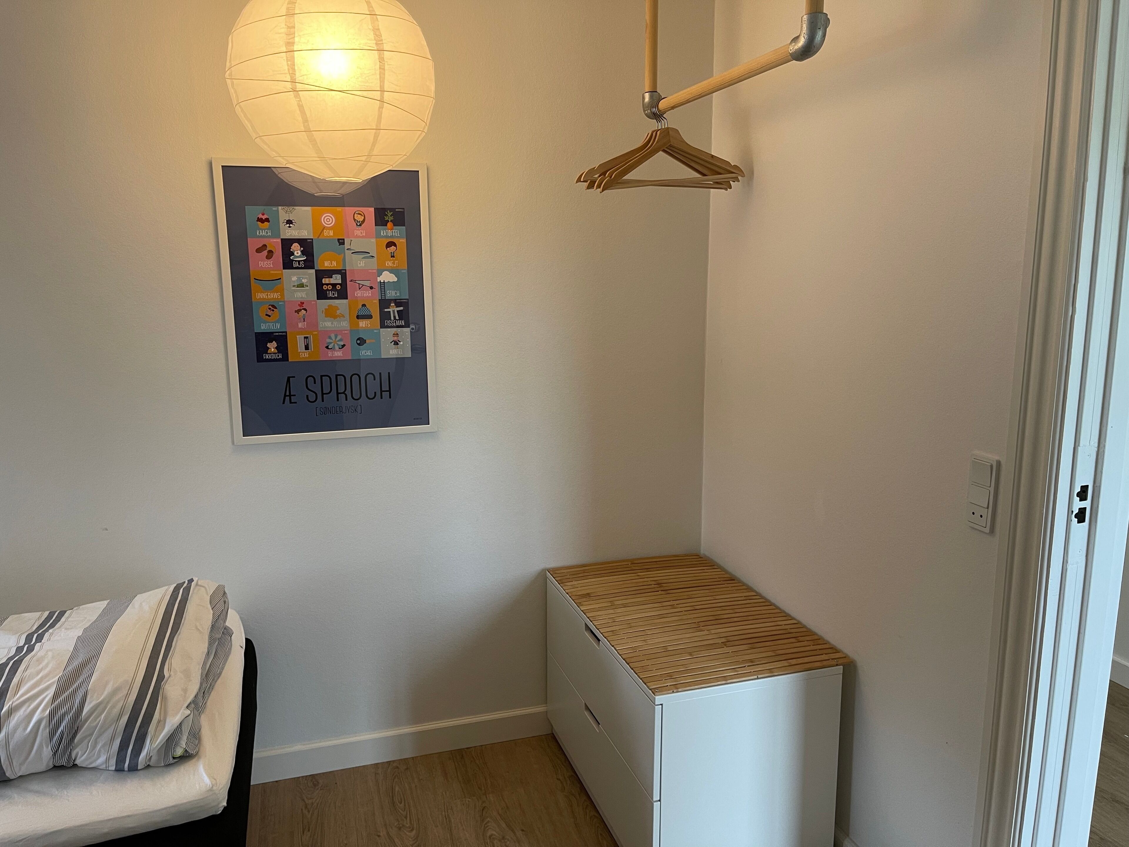 A bedroom with a bed, a small white cabinet, a hanging light fixture, and a poster on the wall.