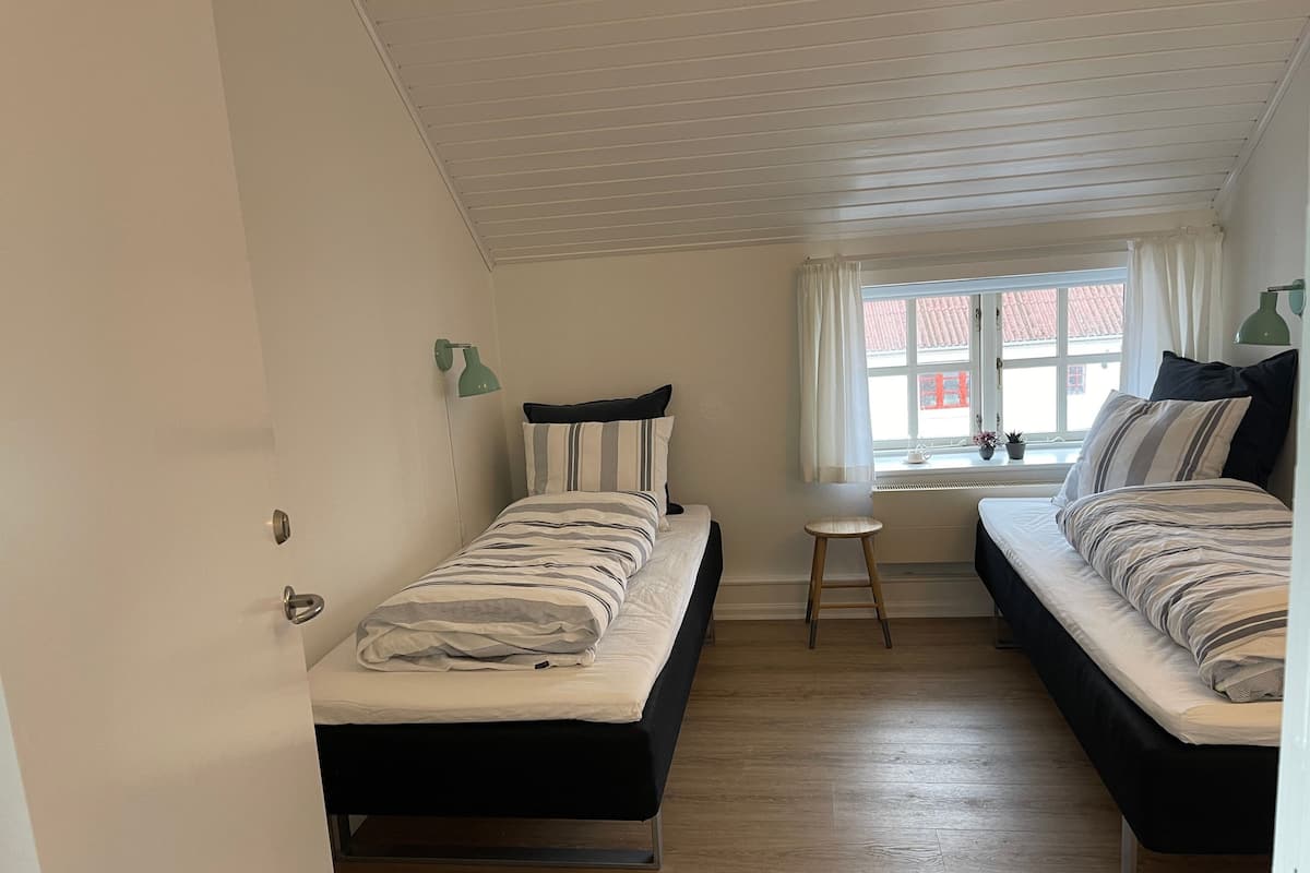Una habitación pequeña y bien iluminada con dos camas, una ventana con cortinas, un taburete y una lámpara de pared.