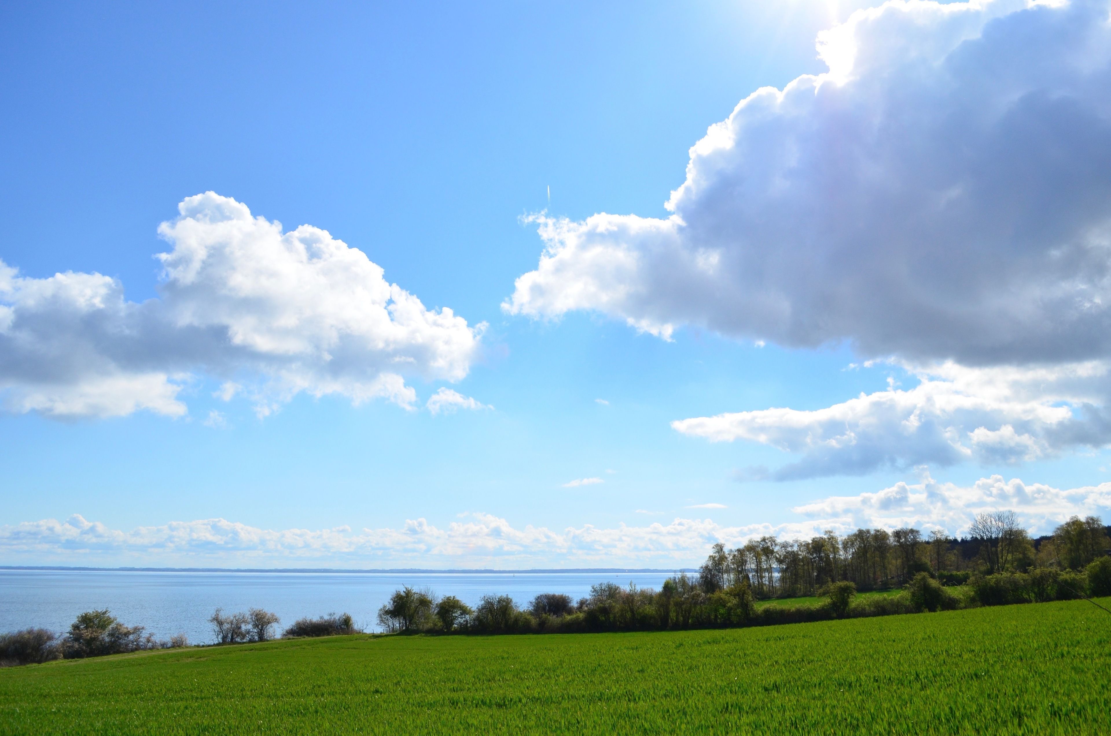 A clear sky with large clouds, a green field, and a body of water in the distance.