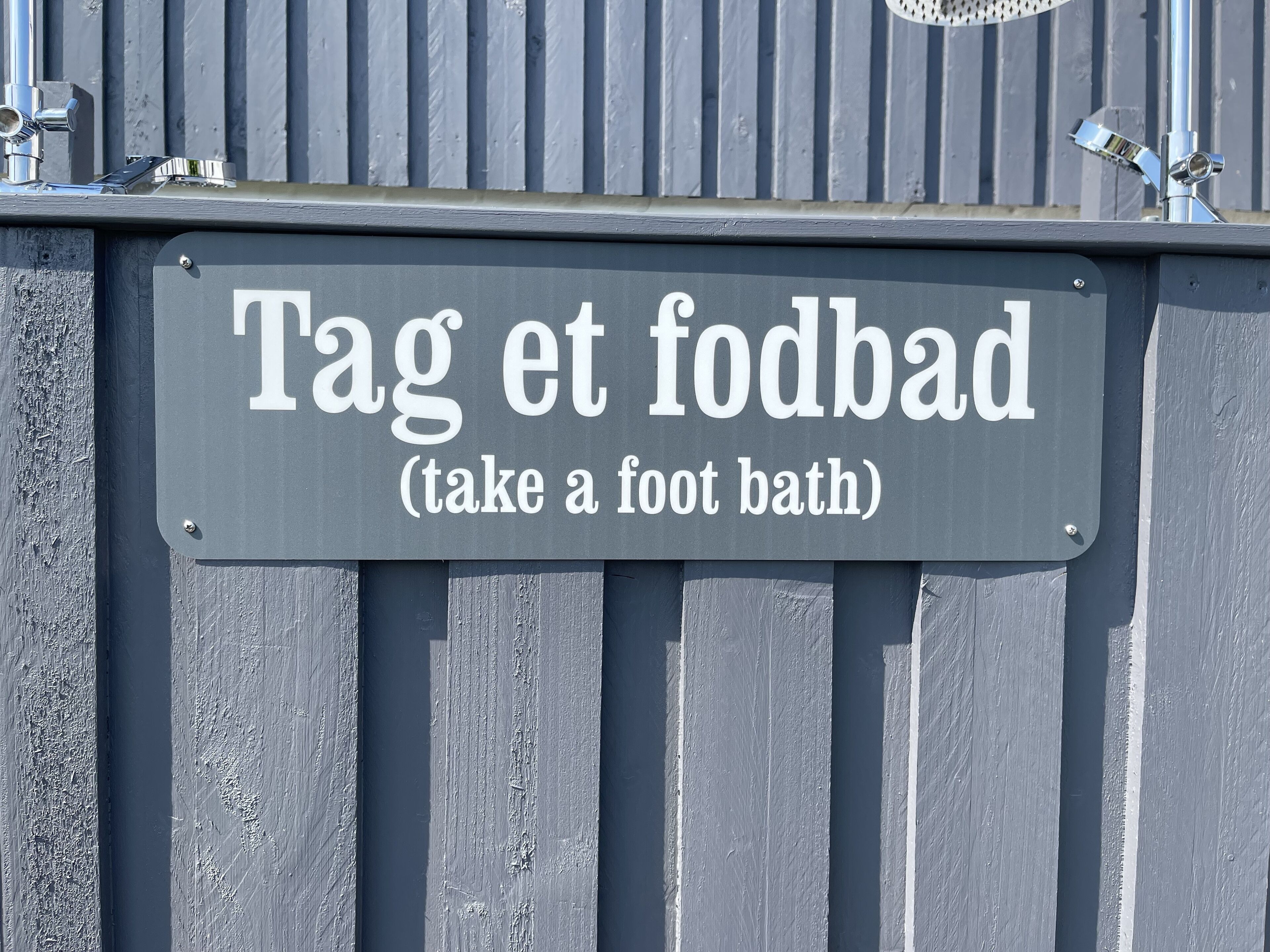 A sign with the text "Tag et fodbad (take a foot bath)" mounted on a grey wall.