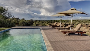 Une piscine à l’eau claire, entourée d’une terrasse en bois. On y trouve des chaises longues et un grand parasol qui offre de l’ombre.