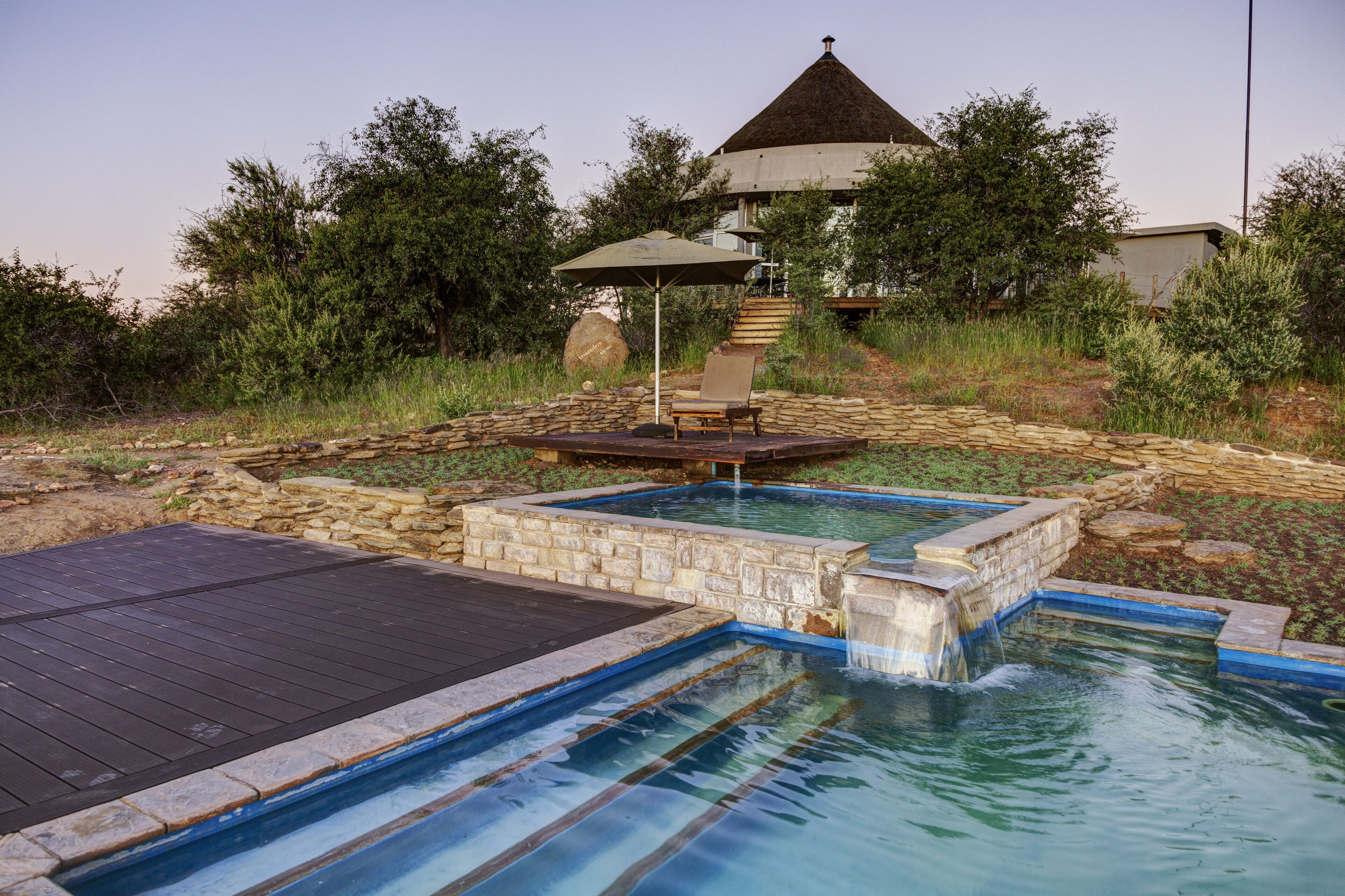 A swimming pool with a wooden deck and stone walls, surrounded by greenery and a gazebo.