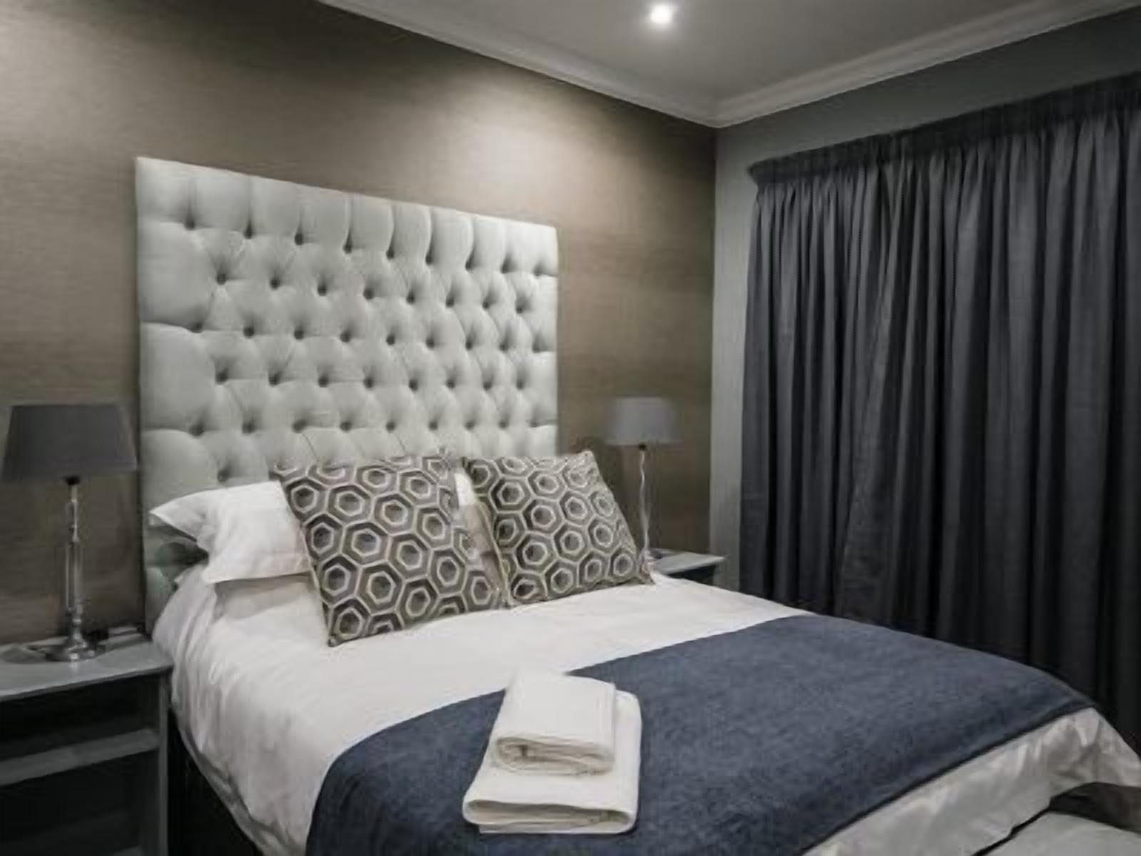 Un lit bien fait, avec une tête de lit capitonnée, deux coussins à motifs, une serviette pliée et une couverture bleu foncé.