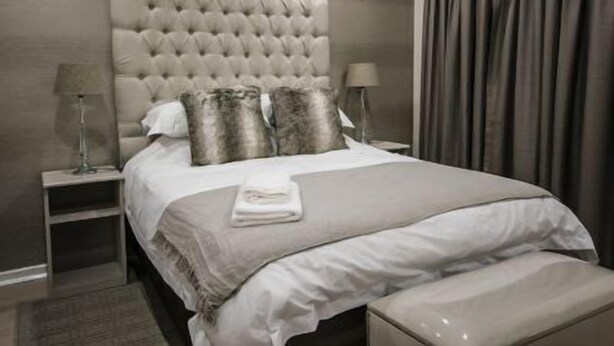A modern bedroom with a tufted headboard, a neatly made bed, a bedside table with a lamp, and a large window with curtains. - The Royal Guest House (Klerksdorp)