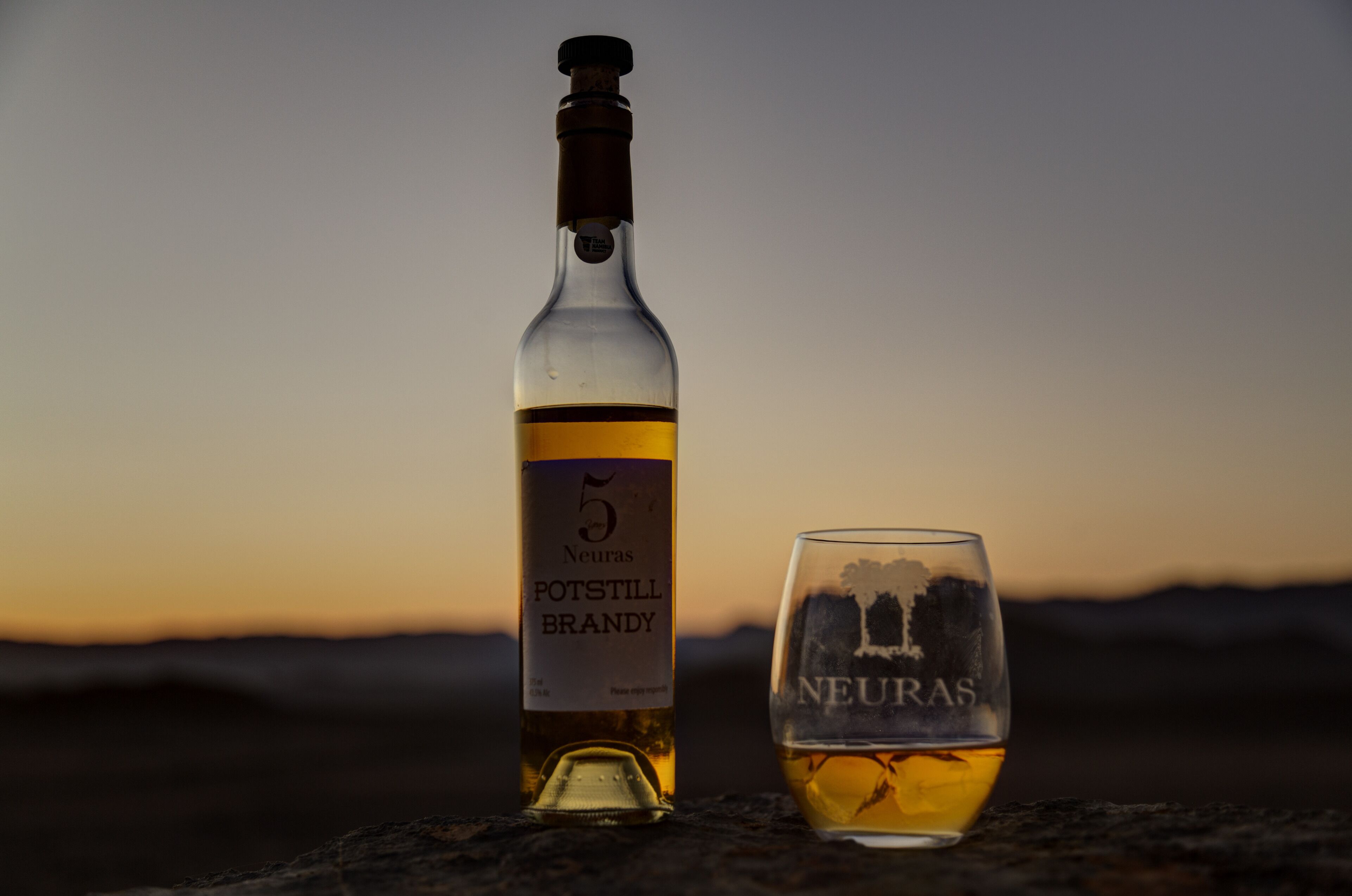Uma garrafa de Brandy Neuras Potstill e um copo sobre uma superfície de pedra, com o pôr do sol ao fundo.