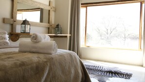 A bedroom with a large bed, a window with curtains, a wooden cross, and a shelf with towels and lanterns. - Neuras Wine & Wildlife Estate (Sesriem)