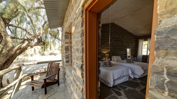 A stone building with a balcony, a wooden bench, and a bedroom with two beds. - Neuras Wine & Wildlife Estate (Sesriem)