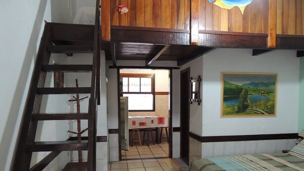 A bedroom with a wooden staircase, a bed, a painting, and a view of a room with a window. - Patricio's Chalés (Ilhabela)