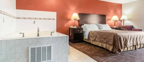 Habitación de hotel con una cama grande, dos mesitas de noche con lámparas, un baño con tocador y lavabo, y una alfombra estampada.
