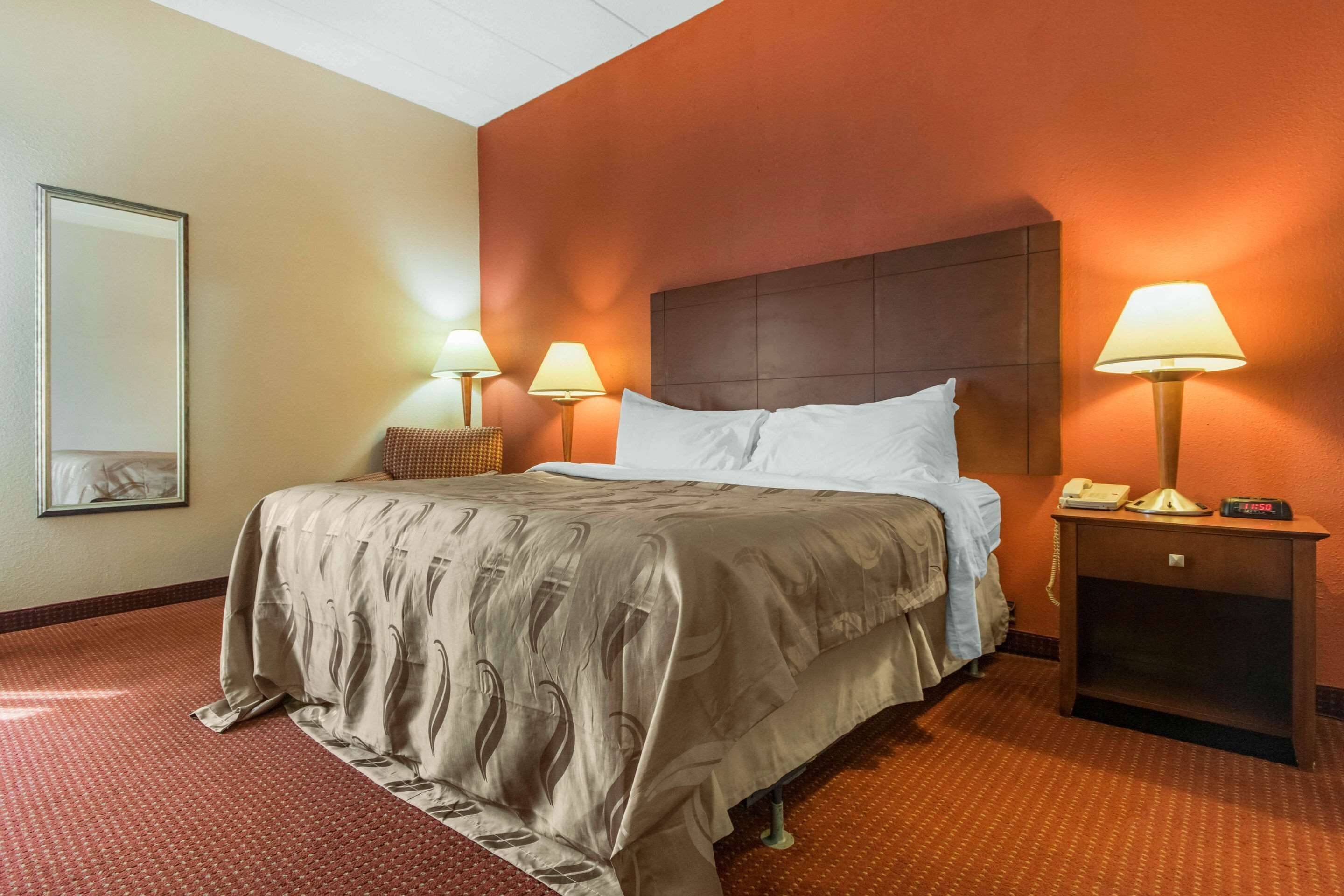 Ein Hotelzimmer mit einem großen Bett, zwei Nachttischlampen, einem Nachttisch mit Telefon und digitaler Uhr sowie einem gerahmten Bild an der Wand.