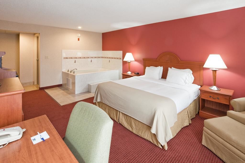 A hotel room with a bed, two bedside tables with lamps, a desk with a telephone, a chair, a small table, and a bathroom with a bathtub and sink.
