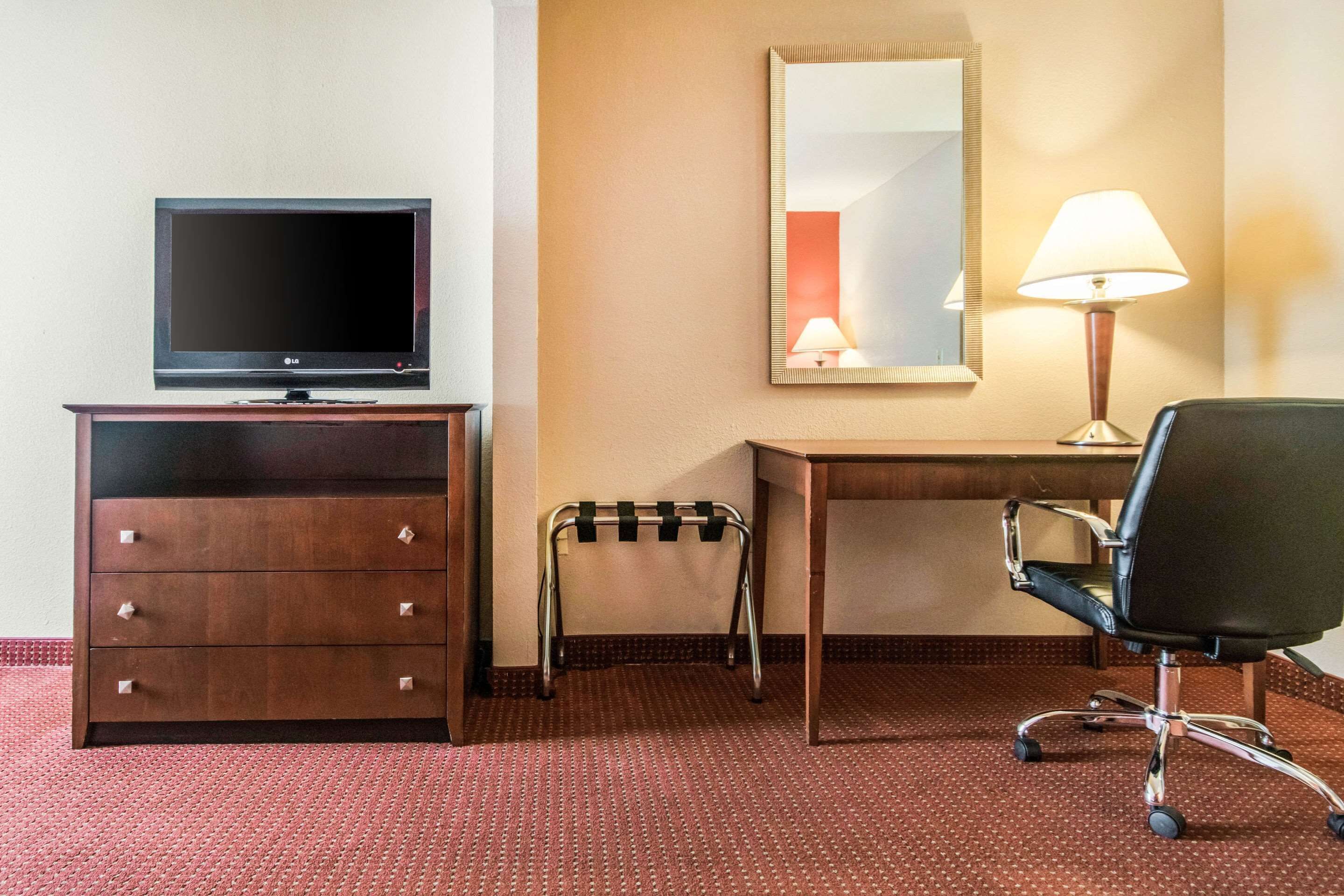 A hotel room with a wooden desk, a black office chair, a television on a wooden dresser, a small stool, a mirror, and a lamp.