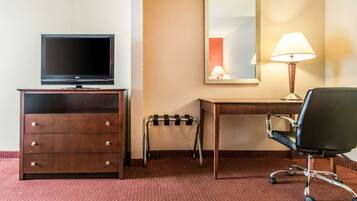 A hotel room with a wooden desk, a black office chair, a television on a wooden dresser, a small stool, a mirror, and a lamp.